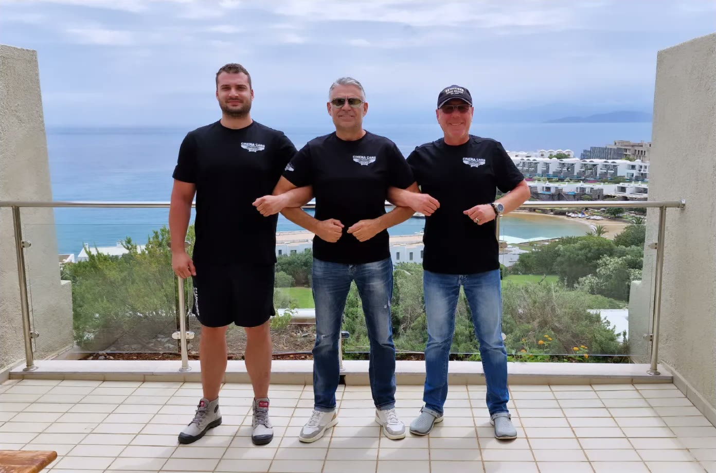 Three members of the Cinema Cars crew in branded T-shirts on a terrace above the sea